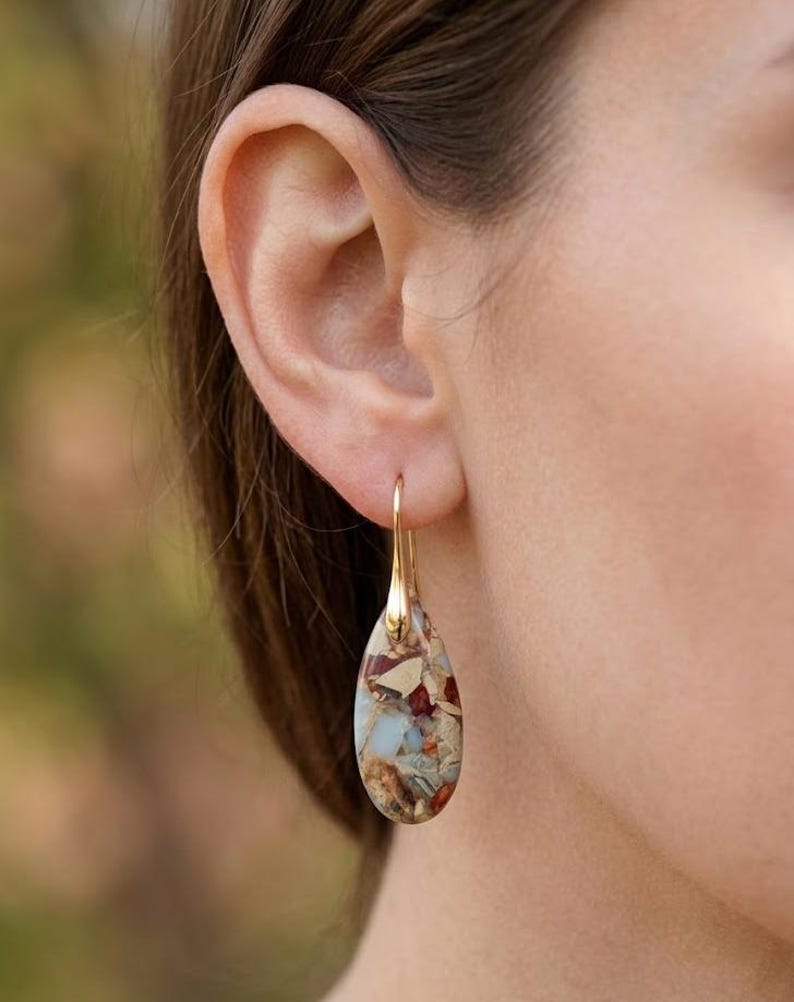 May include: A gold-colored earring with a teardrop-shaped stone. The stone features a mosaic of colors, including beige, red, and blue. The earring is hanging from a person's ear.