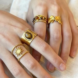 May include: A collection of four gold-toned rings worn on fingers. One ring features a central oval tiger's eye stone, another displays a sunburst motif, a third has three small round stones, and the fourth has a textured, organic leaf-like design.