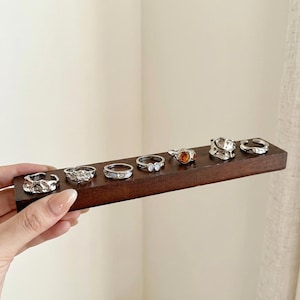 May include: A collection of silver rings displayed on a dark brown wooden holder. The rings feature various designs, including textured, gemstone, and minimalist styles. The rings are held by a person.
