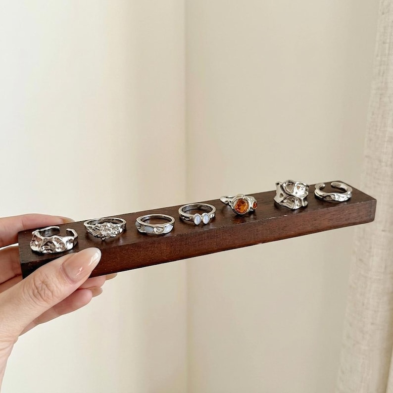 May include: A collection of silver rings displayed on a dark brown wooden holder. The rings feature various designs, including textured, gemstone, and minimalist styles. The rings are held by a person.