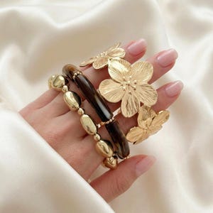 May include: A selection of gold-coloured jewellery, including a beaded bracelet, a dark brown and gold bracelet, and flower-shaped rings. The jewellery is displayed on a white satin surface.