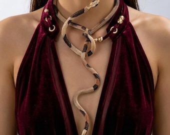 May include: A long, flexible snake-shaped necklace featuring a textured, beige, black, and orange patterned body. The snake wraps around the neck and hangs down the chest, with gold-toned metal accents at the head and tail ends of the piece.