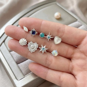 May include: A collection of silver stud earrings in various designs, including a seashell, heart, starburst, and gemstone shapes. The earrings feature opalescent, blue, and clear stones. A pearl is visible in the background.