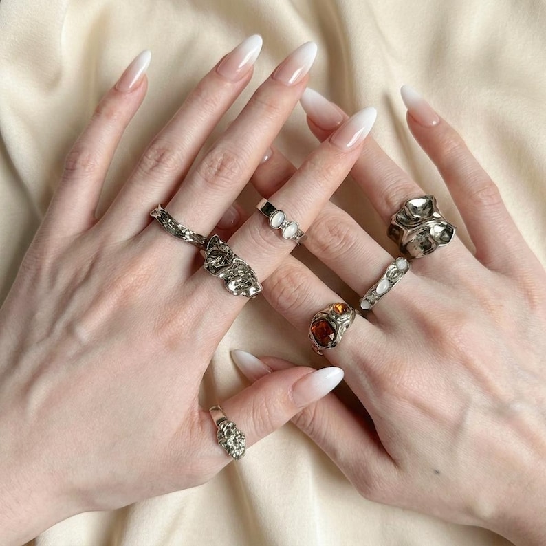 May include: A collection of silver rings, each with unique designs, including a dog-shaped ring, rings with gemstones, and textured bands. The rings are displayed on a hand with long, white-tipped nails, set against a neutral background.