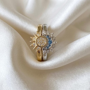 May include: Two stackable rings, one gold and one silver, each featuring a celestial design. The gold ring has a sunburst with a central circle of small, clear stones. The silver ring has a crescent moon with blue stones and clear stones.