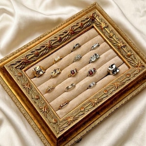 May include: A collection of rings displayed in a decorative, gold-framed jewelry tray. The rings feature various designs, including gemstones and intricate metalwork. The tray has a beige velvet interior, and the frame has ornate detailing.