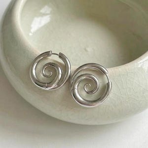 Puede incluir: Un par de pendientes de aro plateados con un diseño en espiral. Los pendientes son circulares y tienen un acabado liso y pulido. Se colocan sobre un fondo de cerámica verde claro.