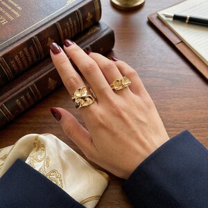 May include: Two gold-colored rings with textured, organic designs are displayed on a hand with dark red nail polish. The rings are set against a backdrop of a wooden surface, books, and a notebook.