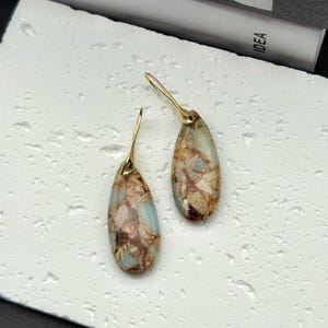 May include: A pair of teardrop-shaped earrings with gold-colored hooks. The earrings feature a marbled pattern in shades of brown, beige, and blue-green. The earrings are displayed on a textured white surface.