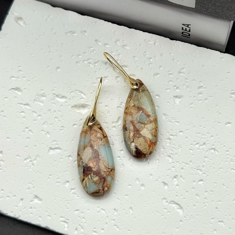 May include: A pair of teardrop-shaped earrings with gold-colored hooks. The earrings feature a marbled pattern in shades of brown, beige, and blue-green. The earrings are displayed on a textured white surface.