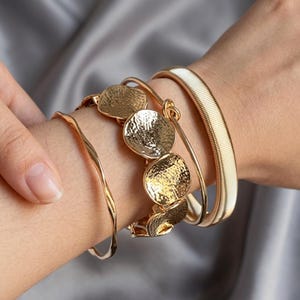 May include: A set of three gold-toned bracelets worn together. One is a thin, smooth, wavy wire bangle. The centre piece is a link bracelet featuring multiple circular, hammered-texture gold discs. The third is a wide, flexible, ribbed metal band.