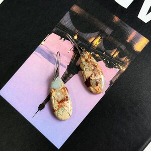 May include: A pair of dangle earrings with teardrop-shaped, multi-colored stone pendants. The stones feature shades of beige, brown, and blue-green. The earrings have silver-colored, curved ear wires. The earrings are displayed on a postcard with a picture of the Eiffel Tower.