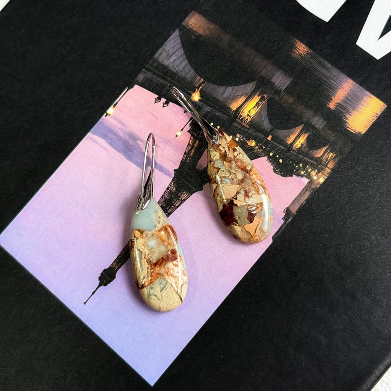 May include: A pair of dangle earrings with teardrop-shaped, multi-colored stone pendants. The stones feature shades of beige, brown, and blue-green. The earrings have silver-colored, curved ear wires. The earrings are displayed on a postcard with a picture of the Eiffel Tower.