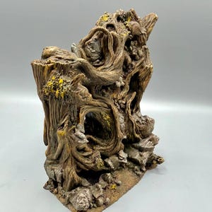 Op de afbeelding: Een gedetailleerde, bruine hars boomstronk met ingewikkelde wortel- en takdetails. De stronk heeft een uitgehold gedeelte en is geaccentueerd met gele en groene vlekken. De basis is getextureerd met kleine rotsen, wat een naturalistische uitstraling creëert.