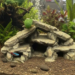 May include: An artificial rock formation for an aquarium, designed to resemble a cave. The rock is a mix of grey and tan, creating a natural appearance. A small, brightly coloured fish swims nearby. The aquarium also contains plants and gravel.