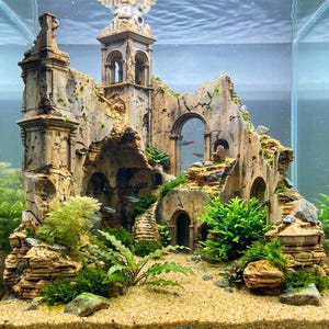 May include: An aquarium ornament depicting a weathered, ruined stone cathedral with arched doorways, a tall tower, and crumbling walls. The structure is surrounded by green aquatic plants and small neon tetra fish swimming through the ruins.