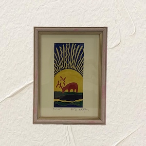 Puede incluir: Obra de arte enmarcada que representa una escena de atardecer con un ciervo. La pintura presenta un sol amarillo con rayos azules, un ciervo rojo y la palabra "Sunset" en la parte inferior. El marco es de color rosa claro.