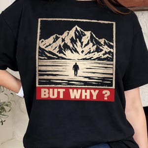 Puede incluir: Camiseta negra con un gráfico de paisaje montañoso, una figura solitaria y el texto "BUT WHY?" en rojo. El diseño incluye una cordillera beige y blanca sobre un fondo negro.