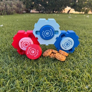 May include: Three brightly coloured dog puzzle toys in red, light blue, and dark blue, with matching circular lids. Each lid has a paw print and the text "PolyPet" and "NoseMe". Several small, round, tan dog treats are also visible.