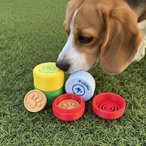 Op de afbeelding: Een beagle snuffelt aan een verzameling kleurrijke bakjes voor dierensnacks. De bakjes zijn geel, groen, rood en wit, met pootafdruk- en logo-ontwerpen. Op een bed van groen gras.