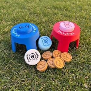 Op de afbeelding: Twee kleine trainingskrukjes voor huisdieren, één blauw en één rood, met de tekst "PolyPet". Verschillende bruine pootafdruk-tokens en twee deksels met de tekst "NoseMe" en "PolyPet" zijn ook zichtbaar. Op groen gras.