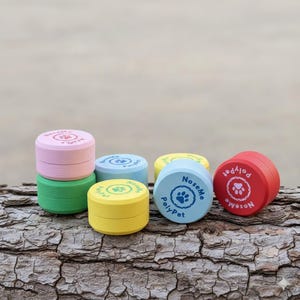 Può includere: Una collezione di contenitori rotondi in legno colorati per dolcetti per animali domestici. Ogni contenitore è di un colore diverso, tra cui rosa, verde, giallo, blu e rosso. I contenitori hanno un design a impronta di zampa e il testo "NoseMe PolyPet".