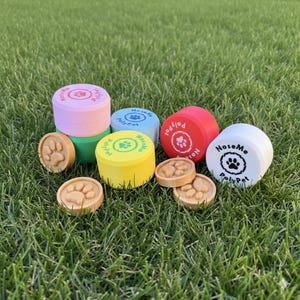 Puede incluir: Una colección de coloridos recipientes para golosinas para perros y golosinas en forma de huellas de patas están dispuestas sobre hierba verde. Los recipientes son cilíndricos, con tapas con el texto "NoseMe PolyPet" y diseños de huellas de patas. Los colores incluyen rosa, verde, azul, amarillo, rojo y blanco.