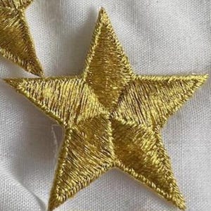 May include: Two gold star-shaped iron-on patches. The stars are five-pointed and have a slightly textured surface.