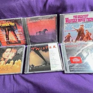 May include: A collection of six CDs featuring movie soundtracks. Titles include "Cowboy Way," "The Horse Whisperer," "The Greatest Western Movie Themes," "Practical Magic," "Twister," and "Original Western Movie Themes." The CDs are in clear plastic cases.