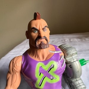 May include: Action figure with a mohawk, purple tank top with a green X, and a silver robotic arm. The figure has a brown beard and a serious expression.