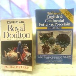 May include: Two books on antiques and collectibles. The book on the left is titled "Official Price Guide to Royal Doulton Sixth Edition" and the book on the right is titled "Warman's English & Continental Pottery & Porcelain 2nd Edition".