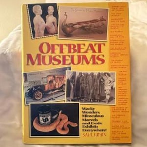 May include: A yellow book titled Offbeat Museums by Saul Rubin. The cover features a collage of photographs including statues, a musical instrument, a vintage car, a group of people, and a mug with a snake, alongside a list of museum names.