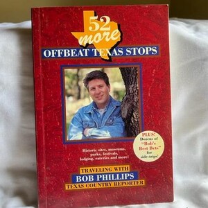 May include: A red book cover with the title "52 more Offbeat Texas Stops" and a photo of a man in a blue denim shirt. The book cover also includes the text "Traveling with Bob Phillips Texas Country Reporter".
