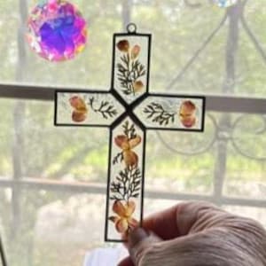 May include: A cross-shaped hanging ornament featuring a black metal frame enclosing clear glass panels. Pressed orange and yellow flowers with green foliage are arranged inside the glass. A small circular metal loop is attached at the top.