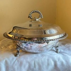 May include: A silver-plated serving dish with a lid and ornate handle. The dish has a rounded shape, rests on three decorative feet, and features a beaded rim. The lid has a small, rectangular emblem. This elegant piece is suitable for serving food.