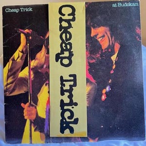 May include: A vintage vinyl record album cover for the band Cheap Trick, featuring live performance shots. The album title, "Cheap Trick at Budokan," is displayed with the band's name in bold, vertical text on a yellow strip.