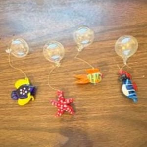 May include: Four decorative ornaments featuring clear glass spheres attached by thin strings to miniature figures: a purple and yellow fish, a red and white polka-dot starfish, an orange and yellow striped fish, and a blue and white seahorse.