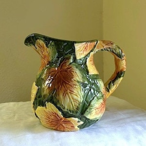 May include: A ceramic pitcher with a green and brown leafy design. The pitcher has a wide mouth and a handle.