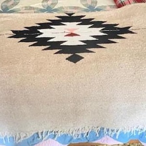 May include: A beige woven blanket with a geometric black, white, and red central design. The blanket has a fringed edge and is displayed on a bed with a patterned bedspread.