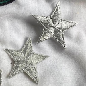 May include: Two silver embroidered star patches. The stars are five-pointed and have a slightly raised texture.