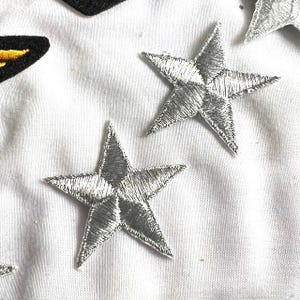 May include: Five silver star-shaped iron-on patches. The stars are a simple design with five points.