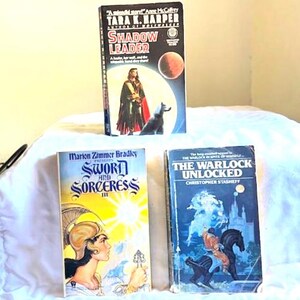 May include: Three paperback books are displayed. The top book is "Shadow Leader" by Tara K. Harper, featuring a figure in a cloak and a dog. Below are "Sword and Sorceress III" and "The Warlock Unlocked".