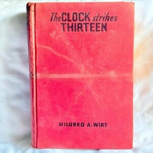 May include: A hardcover book with a bright red cover featuring the title The CLOCK strikes THIRTEEN in black lettering at the top and the author name MILDRED A. WIRT in black lettering at the bottom. The book spine is visible on the left side.