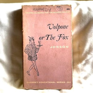 May include: A pink book cover with the title "Volpone or The Fox" by Jonson. The cover features a black and white illustration of a man in a costume holding a tray with a glass of wine.
