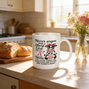 Könnte beinhalten: Weiße Keramik-Tasse mit einer Comic-Kuh und Text. Die Kuh hat eine rosa Schleife. Der Text lautet: „Streite dich niemals mit einem kleinen Mädchen, das einen „A“ in seinem Namen hat...“