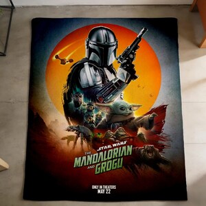 K&ouml;nnte beinhalten: Eine rechteckige Decke mit einer lebendigen Grafik von "The Mandalorian and Grogu". Das Design zeigt den Mandalorianer, Grogu und andere Charaktere vor dem Hintergrund einer orangefarbenen Sonne. Der Text "The Mandalorian and Grogu" ist deutlich sichtbar.