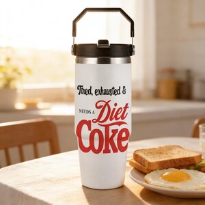May include: A white insulated tumbler with a black handle and lid. The tumbler has the text "Tired, exhausted & NEEDS A Diet Coke" in black and red lettering. The tumbler is on a wooden table next to a plate of food.