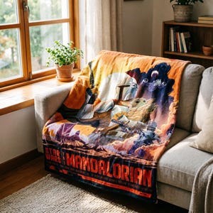K&ouml;nnte beinhalten: Eine Star Wars: The Mandalorian-Decke liegt auf einem grauen Sofa. Die Decke zeigt eine farbenfrohe Grafik des Mandalorianers und Grogu, mit dem Titel der Show unten. Der Hintergrund hat orange, blaue und violette Farbt&ouml;ne.