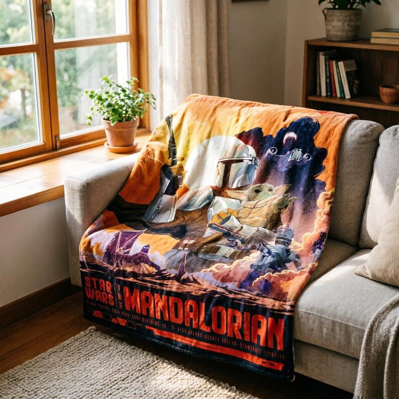 K&ouml;nnte beinhalten: Eine Star Wars: The Mandalorian-Decke liegt auf einem grauen Sofa. Die Decke zeigt eine farbenfrohe Grafik des Mandalorianers und Grogu, mit dem Titel der Show unten. Der Hintergrund hat orange, blaue und violette Farbt&ouml;ne.