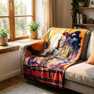 K&ouml;nnte beinhalten: Eine Star Wars: The Mandalorian-Decke liegt auf einem grauen Sofa. Die Decke zeigt eine farbenfrohe Grafik von Mandalorian und Grogu mit den Star Wars- und Mandalorian-Logos in Rot. Die Decke besteht aus weichem Material.